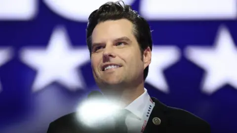 Reuters Wearing a suit with his congressional pin, Matt Gaetz stands at a microphone on a stage with a backdrop of white stars on a blue background. 