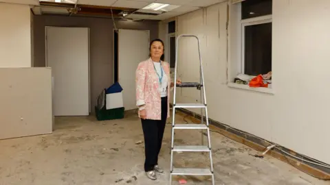 A woman standing in empty room, next to a stepladder. She is wearing dark trousers and a pink and white patterned jacked. The room is unfurnished, and in a state of disrepair, with torn up skirting boards and uncovered and unpainted walls.