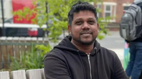Tom MacDougall/BBC Thameshwar Kheran, a man in his mid-30s of South Asian heritage, sitting on one of the benches in the garden.