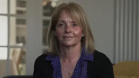 Kirsten Marnie is sat inside a house. She has shoulder length blonde hair and is smiling, looking at the camera. She is wearing two gold necklaces and stud earings in each ear. She is also wearing a pruple patterned blouse and black cardigan.