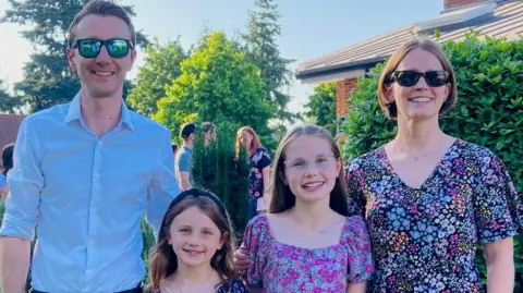 Mike Green The Green family during a party. They are standing outside in a garden. Collette and Mike wear sunglasses. The girls and Collette wear summer dresses. Mike wears a light blue, collared shirt. 