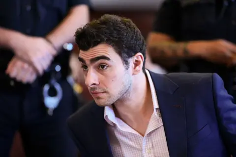 Luigi Mangione appears for a suppression of evidence hearing on 9 December 2025. He wears a pink checkered shirt and navy blue blazer.