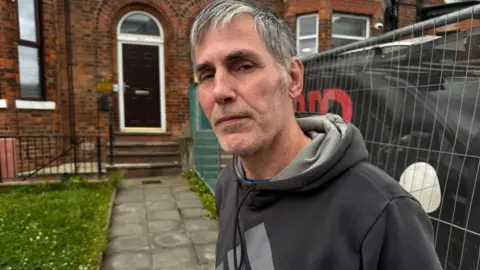 Jonny Humphries/BBC Eddie Aubert, 59, who has grey hair and is wearing a black Adidas branded hoodie, stands outside a terraced house with a work-van parked on the drive way. He is looking into the camera with a serious expression.