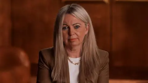 Gavin Hopkins Fiona is photographed against brown background. She is looking stright at the camera. She has long blonde hair and is wearing a brown jacket with a white shirt underneath and a necklace.