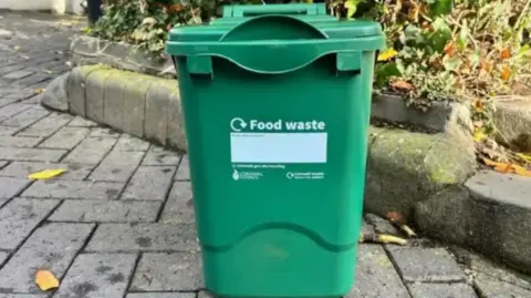 BBC A green food waste caddy on a pavement. It has a recycling logo in white, and the text 'food waste' on the front.