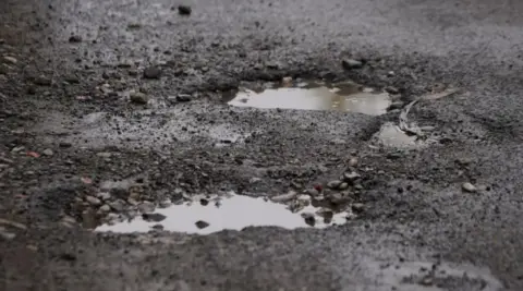 Two potholes in the road next to each other are filled with water. The road surface around them is breaking up.