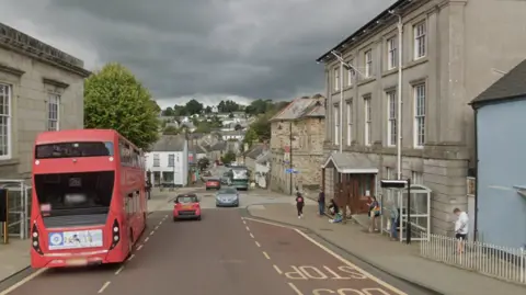 St Nicholas Street stretches into the distance and is lined with stone buildings. There is a bus in the foreground and people waiting at a bus stop opposite.