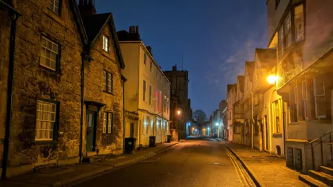 Lucie Johnson A night photo of an empty street in Oxford. There are street lights illuminating the scene. The street is lined with stone houses. 