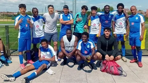 The group of players wearing their blue and white football kits and stood around posing for a photo next to a pitch