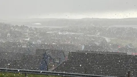 Snow falling across Lerwick in Shetland. Looking out from the top of a hill over lots of houses with black roofs. The sea and hills can be seen in the distance. A dark sky. 