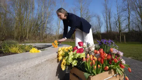 Philippa Rawlinson, a woman wearing glasses and a navy blue jacket and white trousers, is leaning forwards to place a bouquet of yellow flowers on a wall. In the foreground is a box containing a large number of flowers of various colours.