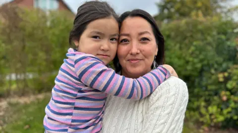 Pilinguaq, who is dressed in a cream jumper with dangly earings, holds her daughter, who is wearing a striped pink, purple and blue top. The daughter is hugging her mother around her shoulders, while in the background there are blurred trees and bushes in front of a house.