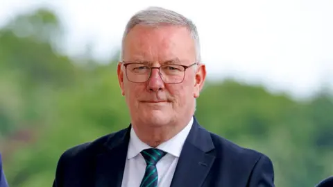 PA Media A man with grey hair wearing thin-framed black glasses.
He is wearing a navy jacket, white shirt and a navy and green diagonal striped tie.