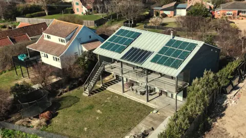 Shaun Whitmore/BBC An aerial image showing a light blue/grey house on which there are solar panels on the roof. It has a garden and next to it is the beach. It is surrounded by other residential properties. 