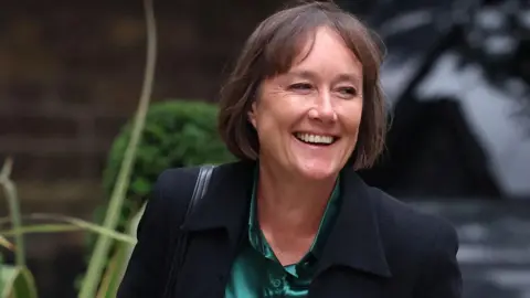 Reuters Jo Stevens in a black suit with a green satin shirt, she is smiling at the camera and holding a red folder