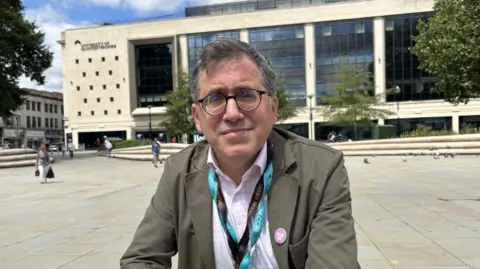 A mid shot of Matthew Reed who's wearing a dark green jacket and light pink shirt. He's got a half smile on his face and is staring at the camera through his glasses. Behind him is Kings Square with the new University of Gloucestershire city campus the back drop, with white stone and black windows.