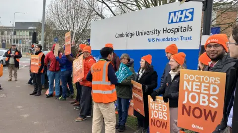 Alex Dunlop/BBC People are stood wearing orange hats and holding placards which say doctors need jobs now. They are stood in front of a blue and white welcome sign outside of a hospital. 