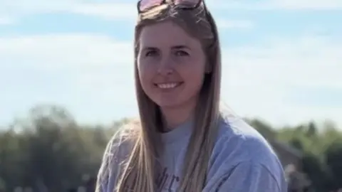 Jennie Logan looking at the camera and smiling. She is wearing a grey sweater and has sunglasses on top of her head.