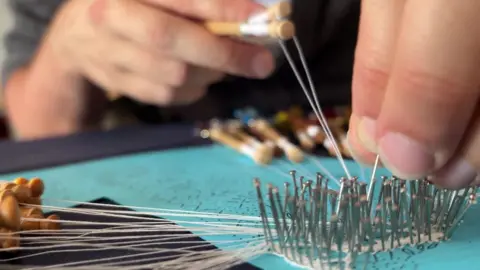 A close-up shot of hands working on bobbin lace, guiding fine white threads through rows of metal pins pinned into a turquoise cushion.