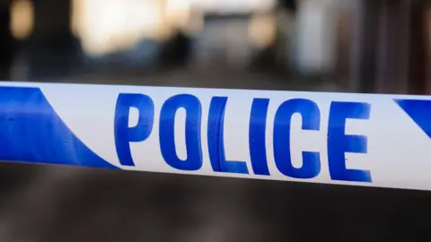 A a close-up of police tape stretched horizontally across the frame. The tape is white with large blue capital letters spelling “POLICE” prominently across the centre.