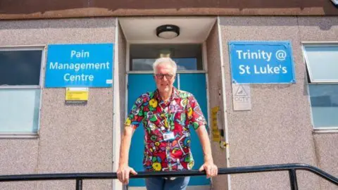 Bradford Teaching Hospitals A man with white curly hair and round glasses wearing a colourful shirt stands outside a clinic