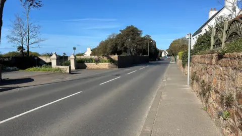 BBC A view of Woodbine corner showing an empty road with houses on one side and a tree. 
