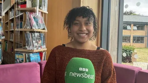 Girl sat on a pink sofa smiling. She has black curly hair tied up with a fringe. She is wearing a dark orange knitwear jumper. to her left there are book shelves behind her. To her right there is a window, you can see a building outside and grass. 