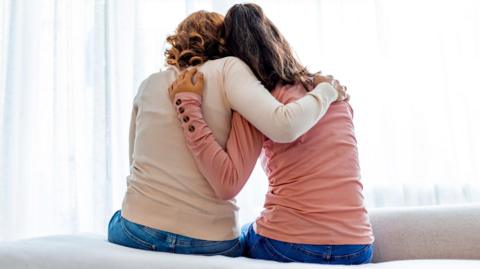 Two women sitting on a bed with their backs to the camera hugging each other. They are wearing jeans and the woman on the right is wearing a peach/pink top and the woman on the left a white jumper