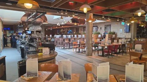 Roger Wood Interior of a restaurant at Gatwick Airport showing rows of tables and chairs, a bar counter, hanging lights and airport facilities. A small number of people can be seen near the bar area while most seating is unused.