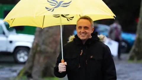 Alex Cole-Hamilton, who has short grey hair, smiles while holding a yellow umbrella