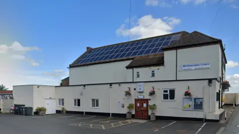 Google A white building with a sign that reads "Birchmeadow Centre". It has wooden double doors and solar panels on the roof, with a blue sky behind it