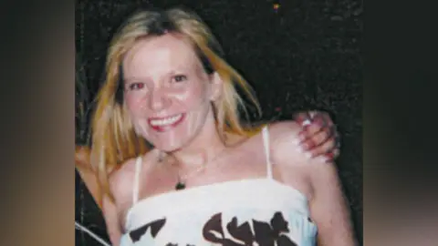 Cathy has long blonde hair and is wearing a white strappy dress with a brown floral pattern and a silver necklace with a black pendant. She is smiling at the camera.