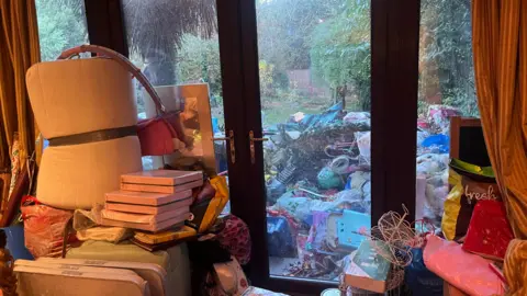 Adrian Harms/BBC A huge amount of rubbish built up in the living room of Nevena Khediri's home in Camberley, Surrey. There is rubbish piled on every surface, and through patio doors a huge amount of additional rubbish can be seen outside.