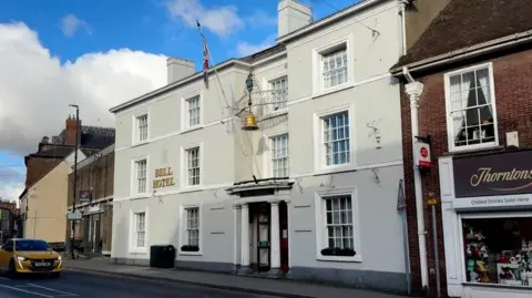 The exterior of the Bell Hotel in Driffield. The Grade II listed building has been painted grey-white and has large sash windows. A golden bell hangs above the main entrance which also has a pole flying the union flag.