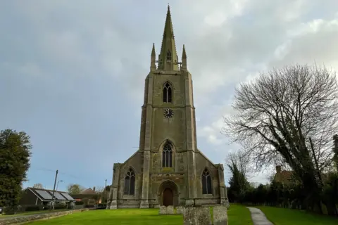 The frontage of a village church with a tall tower and steeple. It stands in pretty grounds with green lawns, gravestones and trees. The tower includes a clock with a black and gold face.