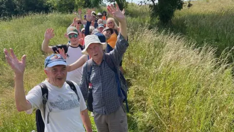 Walkers in a line wave at the camera. They are standing on a footpath running through a field surrounded by grass