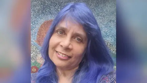 Ness Bobby-Rose Ness Bobby-Rose smiling at the camera with a colourful painting behind her. She has long, purple hair and is wearing a knitted jumper.