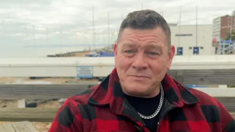 Glyn Busby sits on a bench on the beach, wearing a red and black chequered shirt