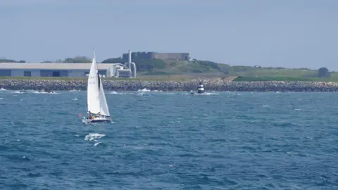BBC Yacht sailing in choppy seas, in the background is an industrial building, a black and white marker and on a grassy hill is Vale Castle.