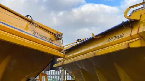 The back of two yellow gritter lorries with the names written on them. On the left is "Grit-asaurus Rex" and on the right is "Grit Britain". The sky can be seen in the background.