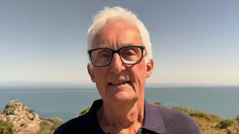 Andrew Mitchell has short white hair and dark-framed glasses and is standing outdoors in bright sunlight. He's wearing a dark navy collared zip-neck top. Behind him, a coastal scene stretches toward the horizon, featuring blue sea, rocky outcrops, and low vegetation in shades of green and brown. The sky is clear and pale blue, and the image is sharply lit by natural daylight.