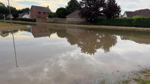 Cambridgeshire Police A road, where a junction would be, has flooded and the water has gone above pavement level. There are houses and trees in the background.