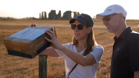 PA Media A woman is holding a cardboard box with a man stood next to her, pointing it at the sun. Behind them is the large Stonehenge circle