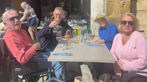 Claire and Neal Hensley Four people sit round a cafe table. One elderly man is in a wheelchair on the left next to Neal, who is holding up a wine glass. Claire is sitting at the front on the right in front of an elderly woman. There are drinks glasses on the table.
