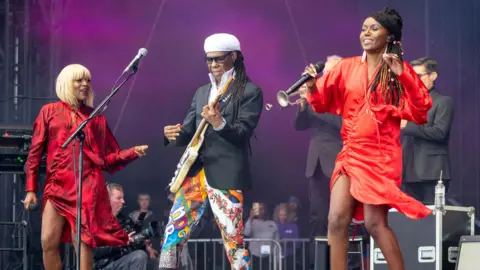 PA Media Nile Rodgers performs onstage at a music festival. He is playing guitar while multi-coloured trousers, a dark jacket and white hat, while two backing singers dance next to him, wearing red dresses.