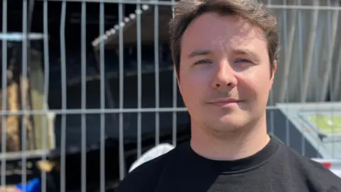 Ryan, a man, with short blonde hair. He is wearing a black T-shirt, smiling for the camera.