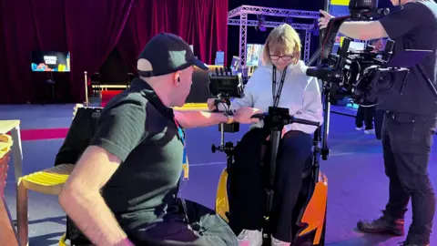 Chris Lynch showing a female student how to use the kit. Jasmine, the student, is sat in a wheelchair learning about the camera's controls.