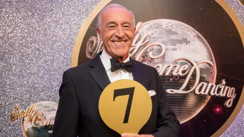 BBC Len Goodman, holding a number seven scorecard, in front of a Strictly Come Dancing logo