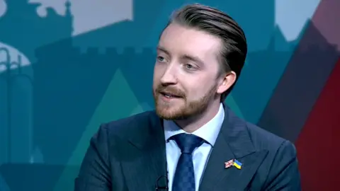 Jaymey McIvor is pictured from the shoulders upwards. He is wearing a blue suit jacket, a blue shirt, and blue tie. Has has two pins on the left side of the jacket, one is a union jack, and the other is a flag of Ukraine. He has brown hair and beard.