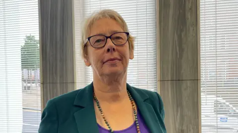 A woman with short blonde hair, glasses, a green jacket, a beaded necklace and a purple top, stands in front of a window with the blinds open.